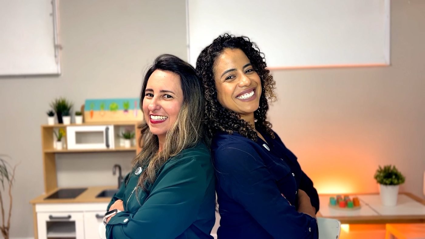 Lisiane e Milene de costas uma para a outra, sorrindo, no estúdio